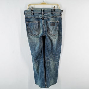 May include: Blue denim jeans with a faded wash, hanging on a hanger. The jeans have a classic five-pocket design, a leather patch on the back, and the Wrangler logo. The jeans are a medium wash with some fading and distressing.