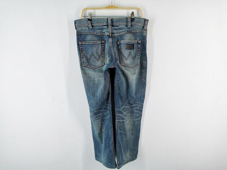 May include: Blue denim jeans with a faded wash, hanging on a hanger. The jeans have a classic five-pocket design, a leather patch on the back, and the Wrangler logo. The jeans are a medium wash with some fading and distressing.