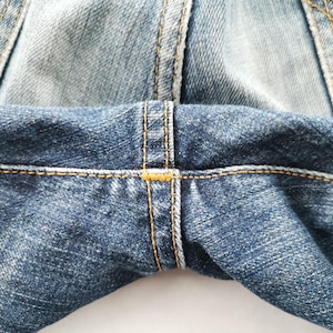 May include: Close-up of denim jeans showing the seam details. The jeans are a medium blue wash with visible stitching in a contrasting yellow-orange thread. The fabric has a textured appearance.