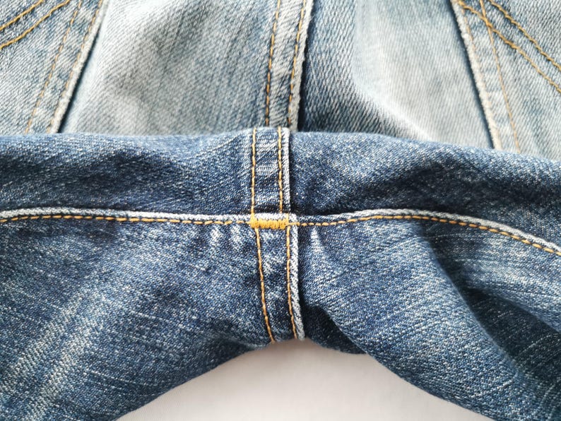 May include: Close-up of denim jeans showing the seam details. The jeans are a medium blue wash with visible stitching in a contrasting yellow-orange thread. The fabric has a textured appearance.