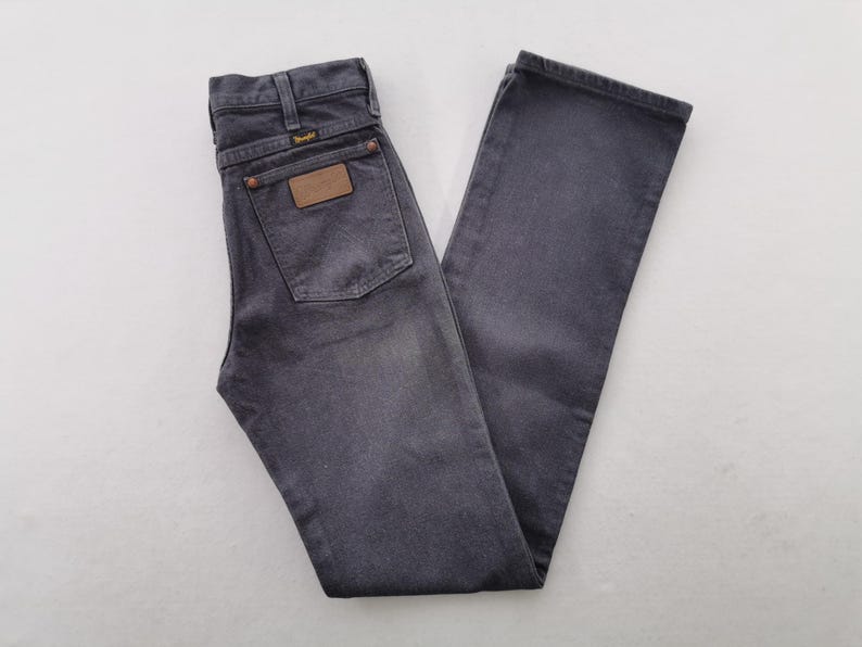 May include: A pair of faded black Wrangler jeans. The jeans have a classic five-pocket design, a brown leather patch on the back, and the Wrangler logo. The jeans are folded and laid flat on a white surface.