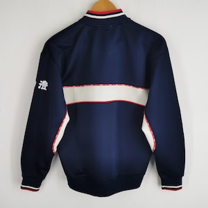 May include: Navy blue track jacket with white and red accents. The jacket features a white stripe across the chest and a small Japanese character on the left sleeve. The collar and cuffs have red and white stripes.