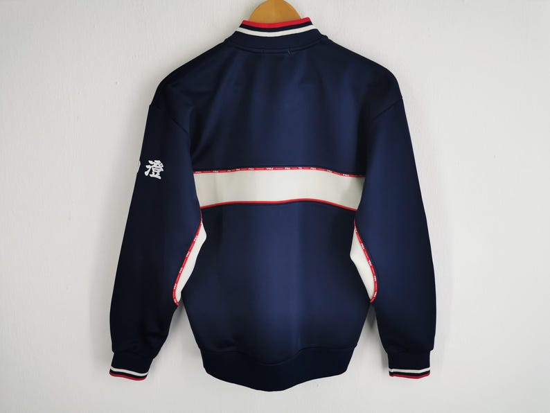 May include: Navy blue track jacket with white and red accents. The jacket features a white stripe across the chest and a small Japanese character on the left sleeve. The collar and cuffs have red and white stripes.