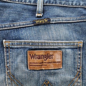 May include: Close-up of blue denim jeans with a Wrangler label on the back pocket. The label is brown with the Wrangler logo and text. The jeans have a classic five-pocket design with visible stitching.