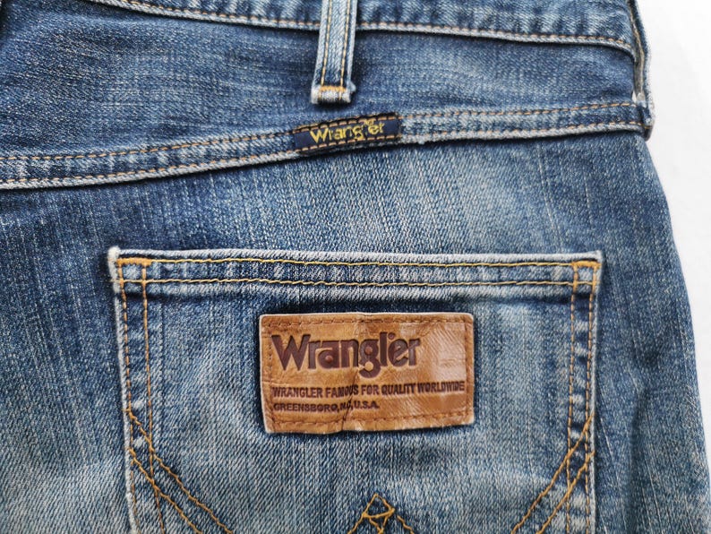 May include: Close-up of blue denim jeans with a Wrangler label on the back pocket. The label is brown with the Wrangler logo and text. The jeans have a classic five-pocket design with visible stitching.