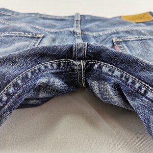 K&ouml;nnte beinhalten: Nahaufnahme einer blauen Jeans, die die Ges&auml;&szlig;taschen und die Nahtdetails zeigt. Die Jeans hat ein abgenutztes Aussehen mit verblichenen Bereichen und wei&szlig;en N&auml;hten. Ein braunes Lederetikett ist sichtbar.