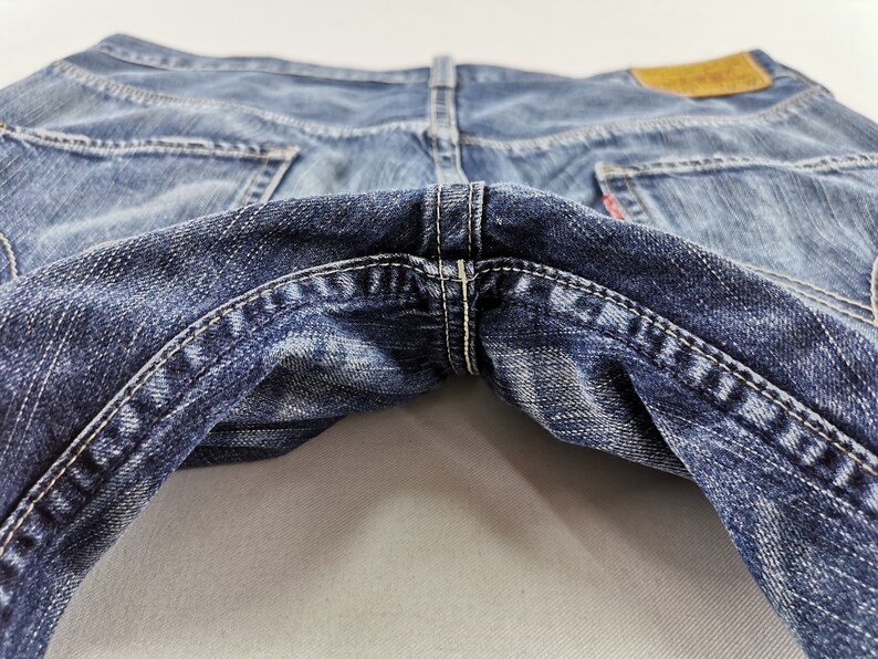 K&ouml;nnte beinhalten: Nahaufnahme einer blauen Jeans, die die Ges&auml;&szlig;taschen und die Nahtdetails zeigt. Die Jeans hat ein abgenutztes Aussehen mit verblichenen Bereichen und wei&szlig;en N&auml;hten. Ein braunes Lederetikett ist sichtbar.