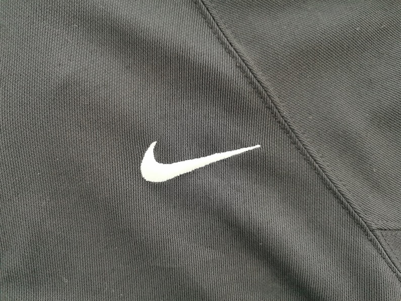 Pu&ograve; includere: Primo piano di tessuto atletico nero con un logo Nike bianco ricamato sul lato sinistro. Il tessuto ha un aspetto strutturato e una cucitura diagonale.
