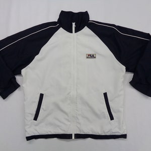 May include: A white and navy blue zip-up track jacket with a stand-up collar. The jacket features navy blue sleeves with white trim and two front pockets. The Fila logo is on the upper left chest.