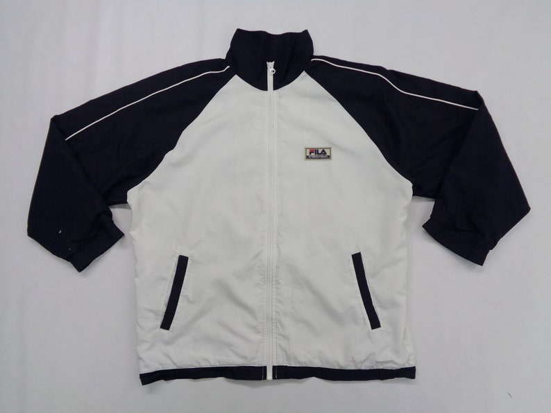 May include: A white and navy blue zip-up track jacket with a stand-up collar. The jacket features navy blue sleeves with white trim and two front pockets. The Fila logo is on the upper left chest.