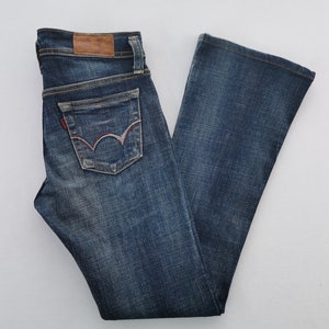 Pu&ograve; includere: Jeans in denim blu scuro con lavaggio sbiadito, una toppa in pelle marrone sulla cintura e una tasca posteriore con un caratteristico motivo ad arco cucito in rosso e bianco. I jeans hanno un taglio a gamba dritta con cuciture dorate visibili.