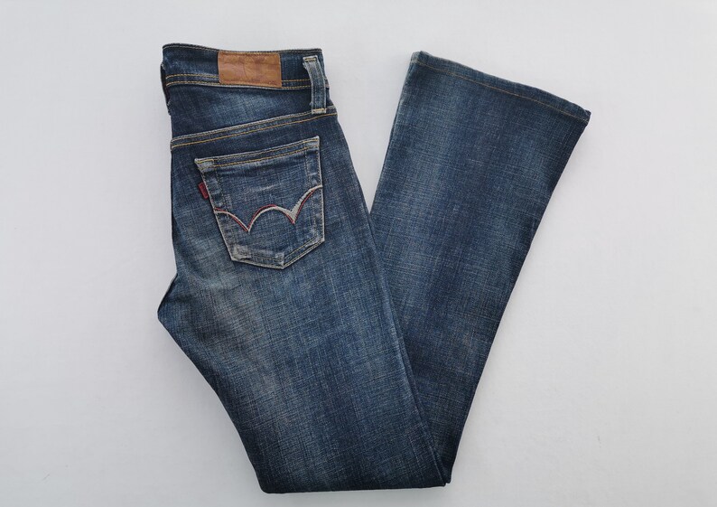 Pu&ograve; includere: Jeans in denim blu scuro con lavaggio sbiadito, una toppa in pelle marrone sulla cintura e una tasca posteriore con un caratteristico motivo ad arco cucito in rosso e bianco. I jeans hanno un taglio a gamba dritta con cuciture dorate visibili.