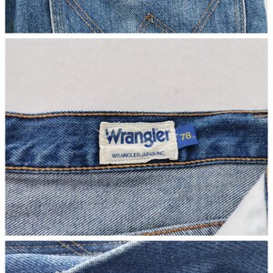 May include: Close-up of blue denim jeans with the Wrangler logo visible on the back pocket and a label. The jeans have a classic design with a button closure and a zipper. The fabric is 100% cotton.