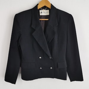 May include: A black double-breasted blazer with silver buttons and a notched lapel. The blazer has two front pockets and is hanging on a wooden hanger. The label reads "Courr&egrave;ges Paris".