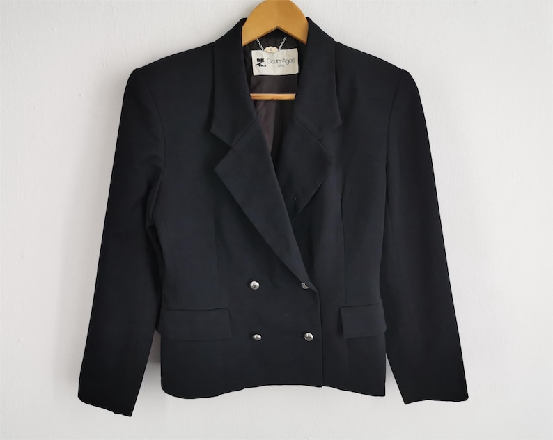 May include: A black double-breasted blazer with silver buttons and a notched lapel. The blazer has two front pockets and is hanging on a wooden hanger. The label reads "Courr&egrave;ges Paris".