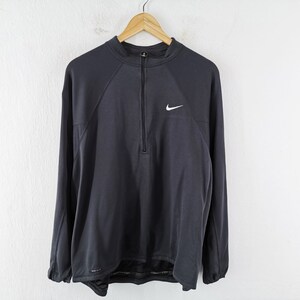 Pu&ograve; includere: Una maglia sportiva Nike a maniche lunghe grigio scuro con chiusura a zip a un quarto e un piccolo logo Nike bianco sul petto. La maglia &egrave; appesa a una gruccia di legno contro un muro bianco.