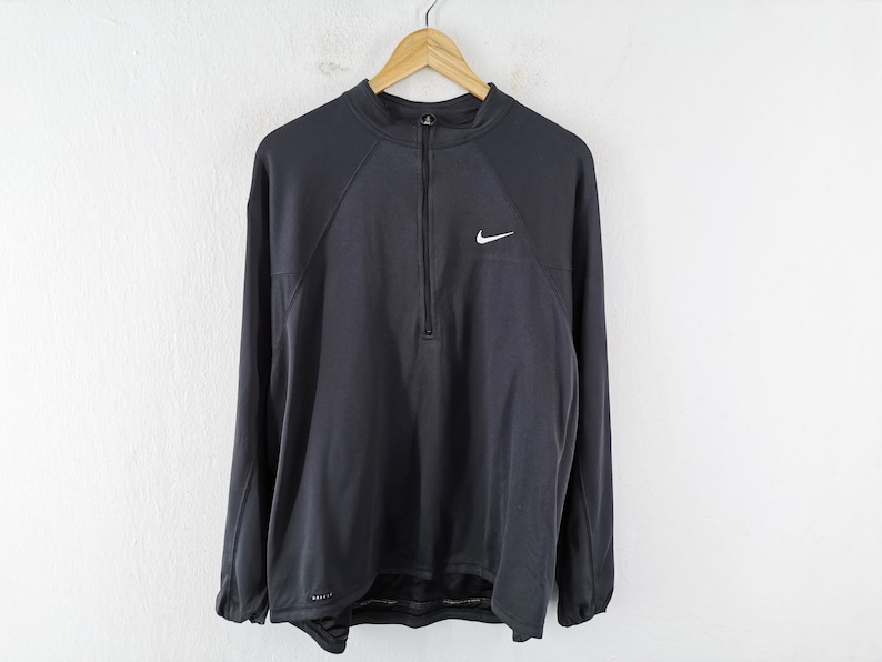 Pu&ograve; includere: Una maglia sportiva Nike a maniche lunghe grigio scuro con chiusura a zip a un quarto e un piccolo logo Nike bianco sul petto. La maglia &egrave; appesa a una gruccia di legno contro un muro bianco.