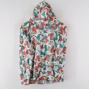 May include: White hooded sweatshirt featuring a repeating pattern of a mermaid with red hair and a teal tail. The design includes other sea creatures and floral elements. The sweatshirt has a drawstring hood and a front pocket.