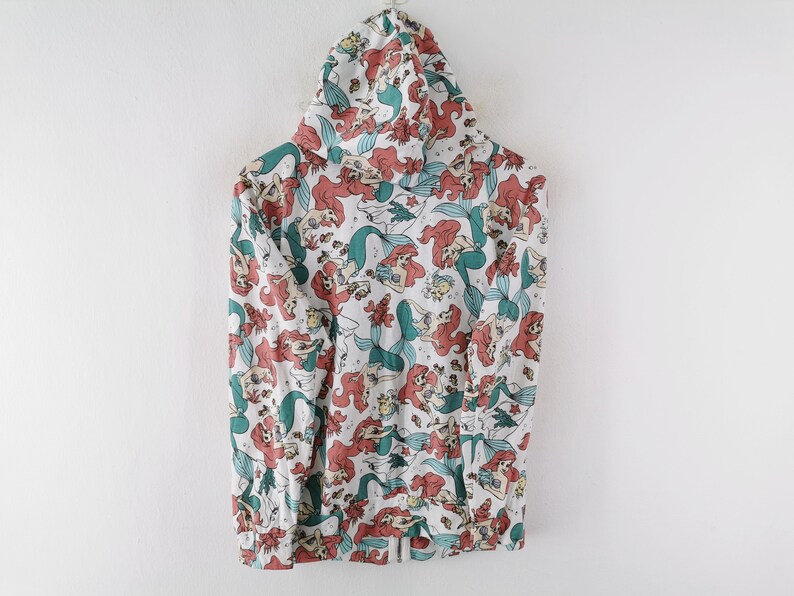 May include: White hooded sweatshirt featuring a repeating pattern of a mermaid with red hair and a teal tail. The design includes other sea creatures and floral elements. The sweatshirt has a drawstring hood and a front pocket.