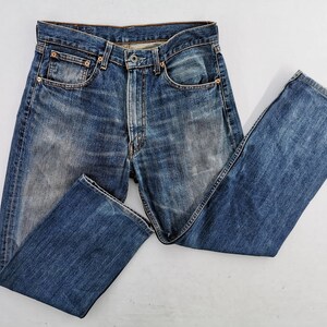 May include: Blue denim jeans with a classic five-pocket design. The jeans have a faded wash and a straight leg cut. The metal rivets and button closure are visible. The jeans are laid flat on a white surface.