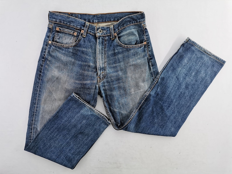 May include: Blue denim jeans with a classic five-pocket design. The jeans have a faded wash and a straight leg cut. The metal rivets and button closure are visible. The jeans are laid flat on a white surface.