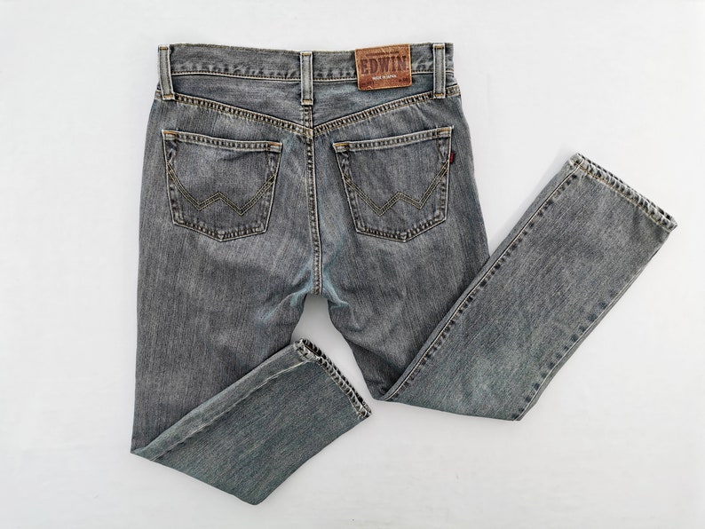 May include: Gray denim jeans with a classic five-pocket design. The jeans feature a faded wash and a leather patch with the brand name "EDWIN" on the back. The back pockets have a distinctive W-shaped stitch.