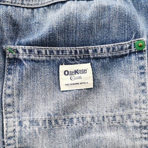 Pu&ograve; includere: Primo piano di tessuto denim blu con una tasca. La tasca presenta un'etichetta bianca con il logo "OshKosh Classic" e il testo "THE GENUINE ARTICLE". La tasca &egrave; fissata con un bottone verde.