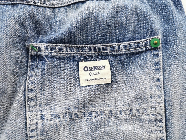 Pu&ograve; includere: Primo piano di tessuto denim blu con una tasca. La tasca presenta un'etichetta bianca con il logo "OshKosh Classic" e il testo "THE GENUINE ARTICLE". La tasca &egrave; fissata con un bottone verde.