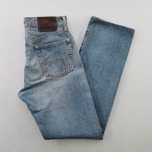 May include: Light blue denim jeans with a classic five-pocket design. The jeans feature a faded wash, a brown leather patch, and a small orange tag. The back pockets have a distinctive stitch pattern. A casual style.