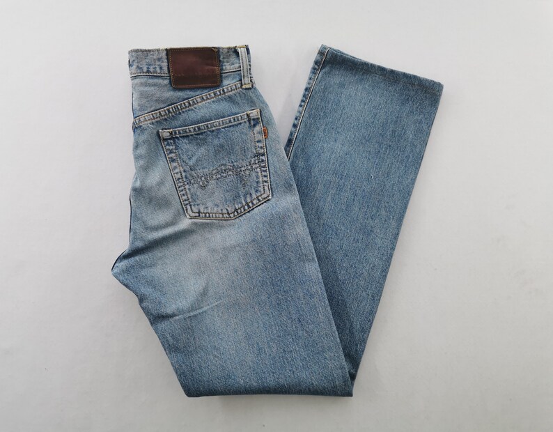 May include: Light blue denim jeans with a classic five-pocket design. The jeans feature a faded wash, a brown leather patch, and a small orange tag. The back pockets have a distinctive stitch pattern. A casual style.