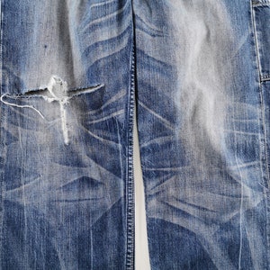 K&ouml;nnte beinhalten: Nahaufnahme einer zerrissenen blauen Jeans aus Denim mit einem gro&szlig;en Riss in Knieh&ouml;he. Die Jeans hat verblasste Bereiche und sichtbare Abnutzungsmuster, die einen Vintage- oder Used-Look zeigen. Der Stoff ist ein mittelschwerer Denim.
