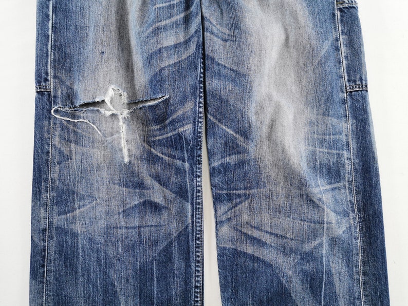 K&ouml;nnte beinhalten: Nahaufnahme einer zerrissenen blauen Jeans aus Denim mit einem gro&szlig;en Riss in Knieh&ouml;he. Die Jeans hat verblasste Bereiche und sichtbare Abnutzungsmuster, die einen Vintage- oder Used-Look zeigen. Der Stoff ist ein mittelschwerer Denim.