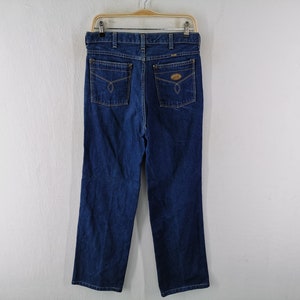 May include: Dark blue denim jeans with a classic five-pocket design. The jeans are hanging on a wooden hanger against a white background. Features include a button and zipper closure, belt loops, and two back pockets with decorative stitching.