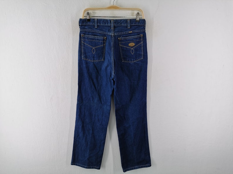 May include: Dark blue denim jeans with a classic five-pocket design. The jeans are hanging on a wooden hanger against a white background. Features include a button and zipper closure, belt loops, and two back pockets with decorative stitching.