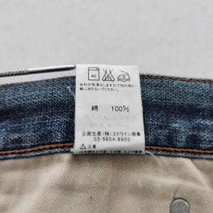 K&ouml;nnte beinhalten: Nahaufnahme eines Jeanskleidungsst&uuml;cks mit einem wei&szlig;en Etikett. Das Etikett zeigt japanischen Text, Pflegesymbole und den Text "100%", der die Materialzusammensetzung angibt. Die N&auml;hte und ein Metallknopf des Kleidungsst&uuml;cks sind sichtbar.