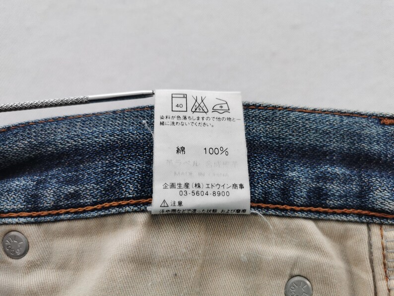 K&ouml;nnte beinhalten: Nahaufnahme eines Jeanskleidungsst&uuml;cks mit einem wei&szlig;en Etikett. Das Etikett zeigt japanischen Text, Pflegesymbole und den Text "100%", der die Materialzusammensetzung angibt. Die N&auml;hte und ein Metallknopf des Kleidungsst&uuml;cks sind sichtbar.