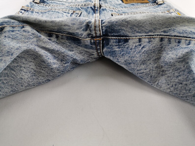 K&ouml;nnte beinhalten: Nahaufnahme einer Jeans aus Acid-Wash-Denim. Die Jeans hat ein hellblaues und wei&szlig;es Marmormuster. Die Ges&auml;&szlig;taschen und der Bund sind sichtbar. Die N&auml;hte sind in einer kontrastierenden orangefarbenen Farbe gehalten.