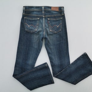 Pu&ograve; includere: Un paio di jeans in denim blu scuro con lavaggio sbiadito, una toppa in pelle marrone sulla cintura posteriore e due tasche posteriori con un caratteristico design a cuciture curve in filo chiaro. I jeans hanno il classico modello a cinque tasche.