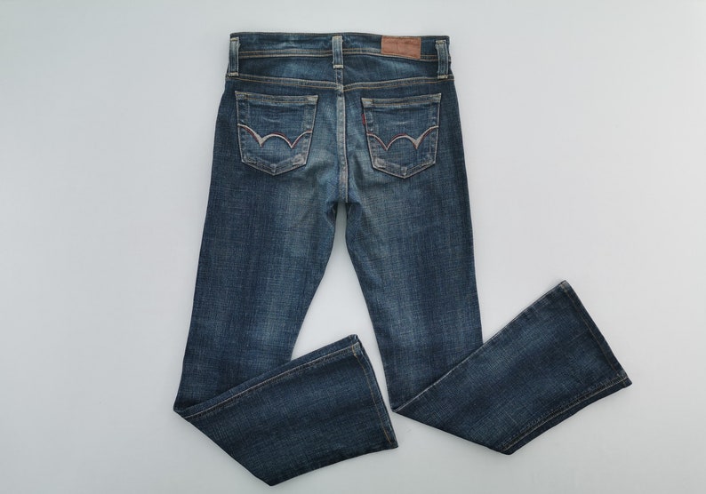 Pu&ograve; includere: Un paio di jeans in denim blu scuro con lavaggio sbiadito, una toppa in pelle marrone sulla cintura posteriore e due tasche posteriori con un caratteristico design a cuciture curve in filo chiaro. I jeans hanno il classico modello a cinque tasche.