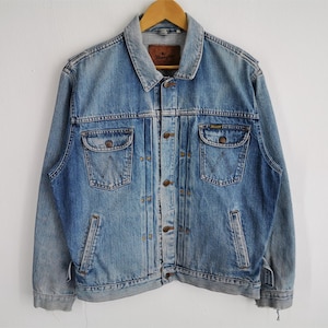 May include: A light blue denim jacket with a button-down front, two chest pockets, and two side pockets. The jacket has a classic collar and a brown leather patch on the back of the collar. The jacket is a vintage Wrangler brand.
