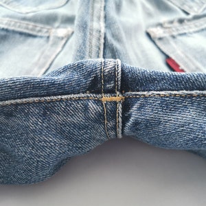 May include: Close-up of light blue denim jeans. The image highlights the stitching details at the crotch and the back pockets. The denim has a worn appearance, with visible seams and a red tag.