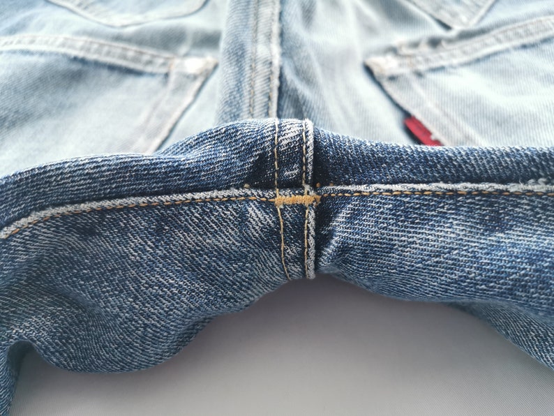 May include: Close-up of light blue denim jeans. The image highlights the stitching details at the crotch and the back pockets. The denim has a worn appearance, with visible seams and a red tag.