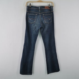 Pu&ograve; includere: Un paio di jeans in denim blu scuro con un classico design a cinque tasche, una toppa in pelle marrone sulla cintura posteriore e cuciture decorative sulle due tasche posteriori. I jeans sono appesi a una gruccia di plastica.