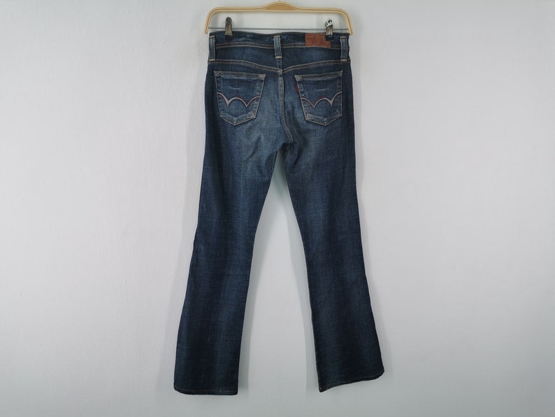 Pu&ograve; includere: Un paio di jeans in denim blu scuro con un classico design a cinque tasche, una toppa in pelle marrone sulla cintura posteriore e cuciture decorative sulle due tasche posteriori. I jeans sono appesi a una gruccia di plastica.