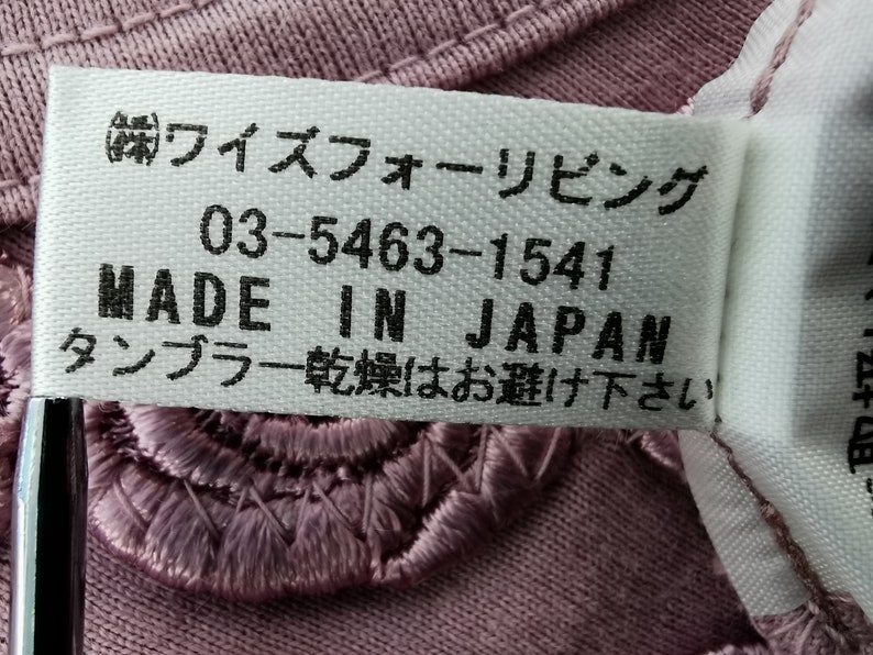 May include: Close-up of a label with Japanese text and the words "MADE IN JAPAN." The label is white with black text and is sewn onto a mauve fabric with embroidered floral details.