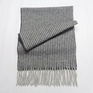 May include: A gray scarf with vertical dark gray stripes and fringe. The scarf is folded over itself, showcasing the striped pattern. The fringe is a lighter shade of gray. The scarf appears to be made of a soft, woven material.
