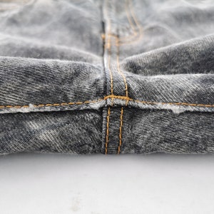 May include: Close-up of the crotch area of a pair of faded black denim jeans. The jeans have orange stitching along the seams and a distressed look.