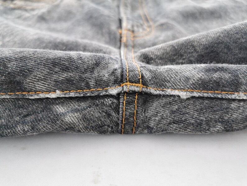 May include: Close-up of the crotch area of a pair of faded black denim jeans. The jeans have orange stitching along the seams and a distressed look.