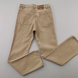 May include: Tan-colored denim jeans with a classic five-pocket design. The jeans feature a brown leather patch on the back waistband and a small red tag on the back pocket. The pants are laid flat, showcasing the details of the stitching and the overall style.