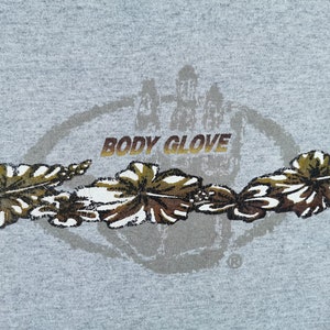 May include: A gray t-shirt featuring the "BODY GLOVE" logo in gold lettering, with a circular design and a handprint graphic. Below the logo are stylized brown and white floral designs.
